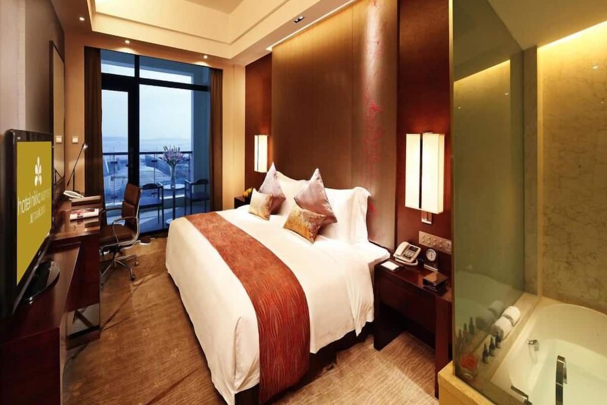 Habitación de hotel con una cama grande, un escritorio, una silla, un teléfono y un baño con paredes de vidrio.