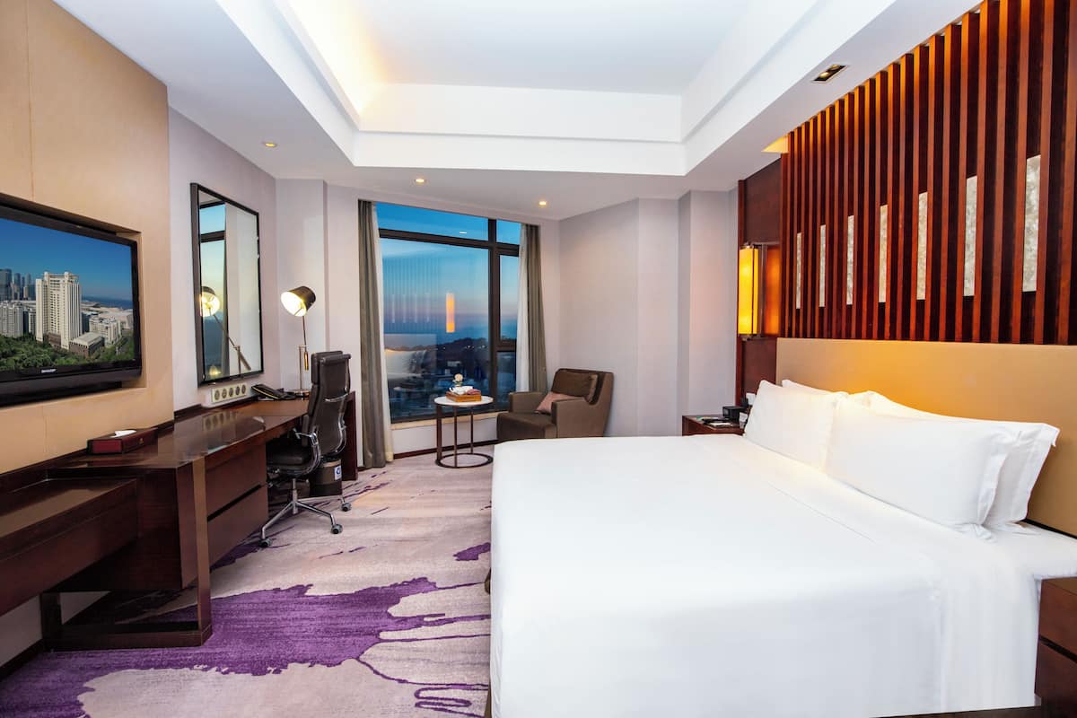 Habitación de hotel con una cama grande, un escritorio, una silla, una mesita y un amplio ventanal con vistas a la ciudad.