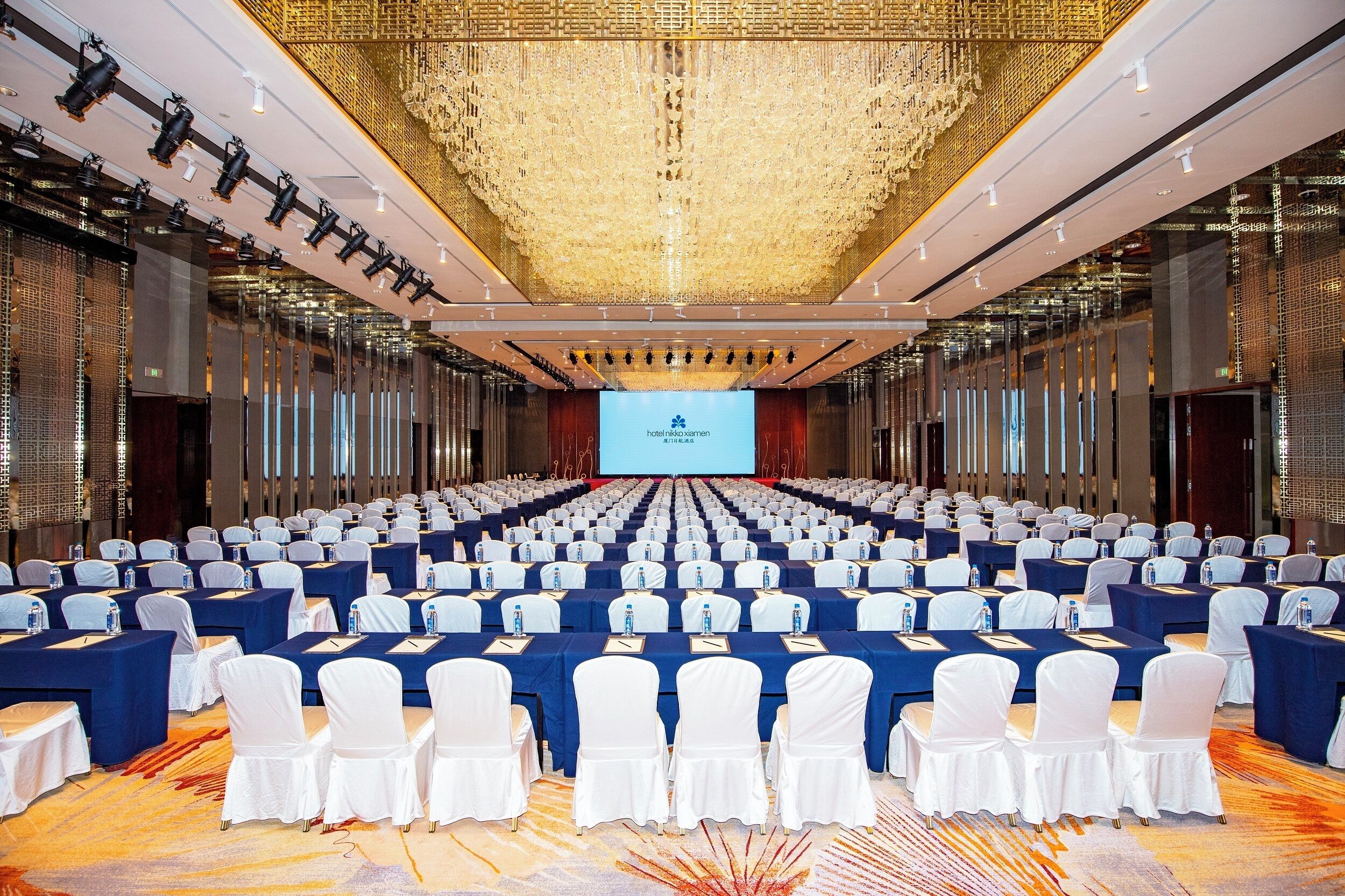 Un amplio salón de conferencias con filas de mesas y sillas, una pantalla grande al frente y un techo decorativo.