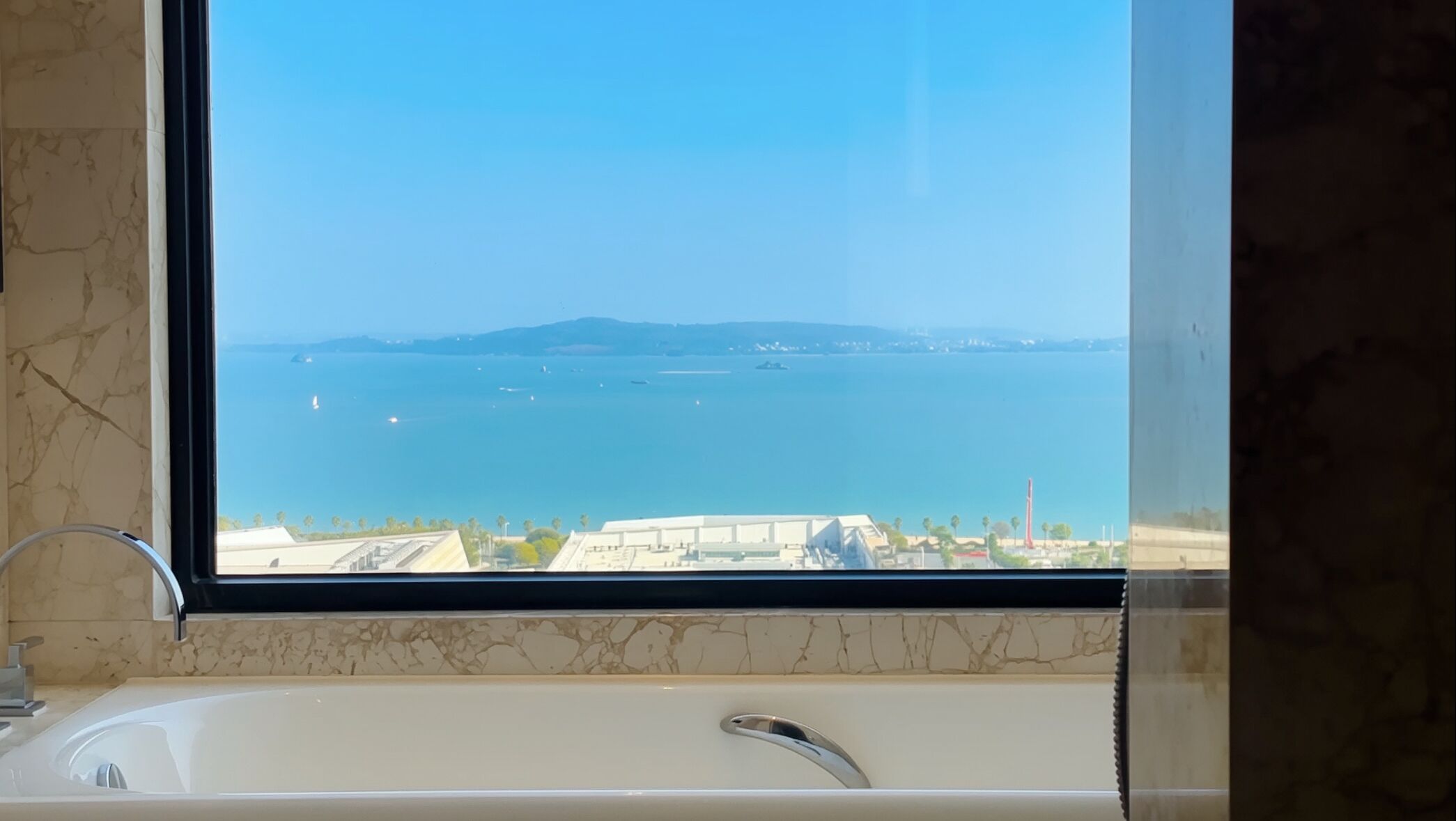 A bathroom with a bathtub, a marble wall, and a window overlooking a body of water with boats.