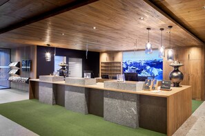 A reception desk with a wooden ceiling, a large screen displaying a snowy mountain landscape, and a green carpeted floor. - Zugspitz Resort (Ehrwald)