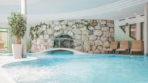 An indoor swimming pool with a stone wall and a metal arch bridge. There are lounge chairs and a potted plant.