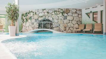 An indoor swimming pool with a stone wall and a metal arch bridge. There are lounge chairs and a potted plant.