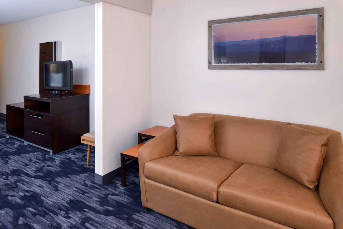 A hotel room with a brown leather sofa, a small wooden table, a TV on a cabinet, and a painting on the wall.
