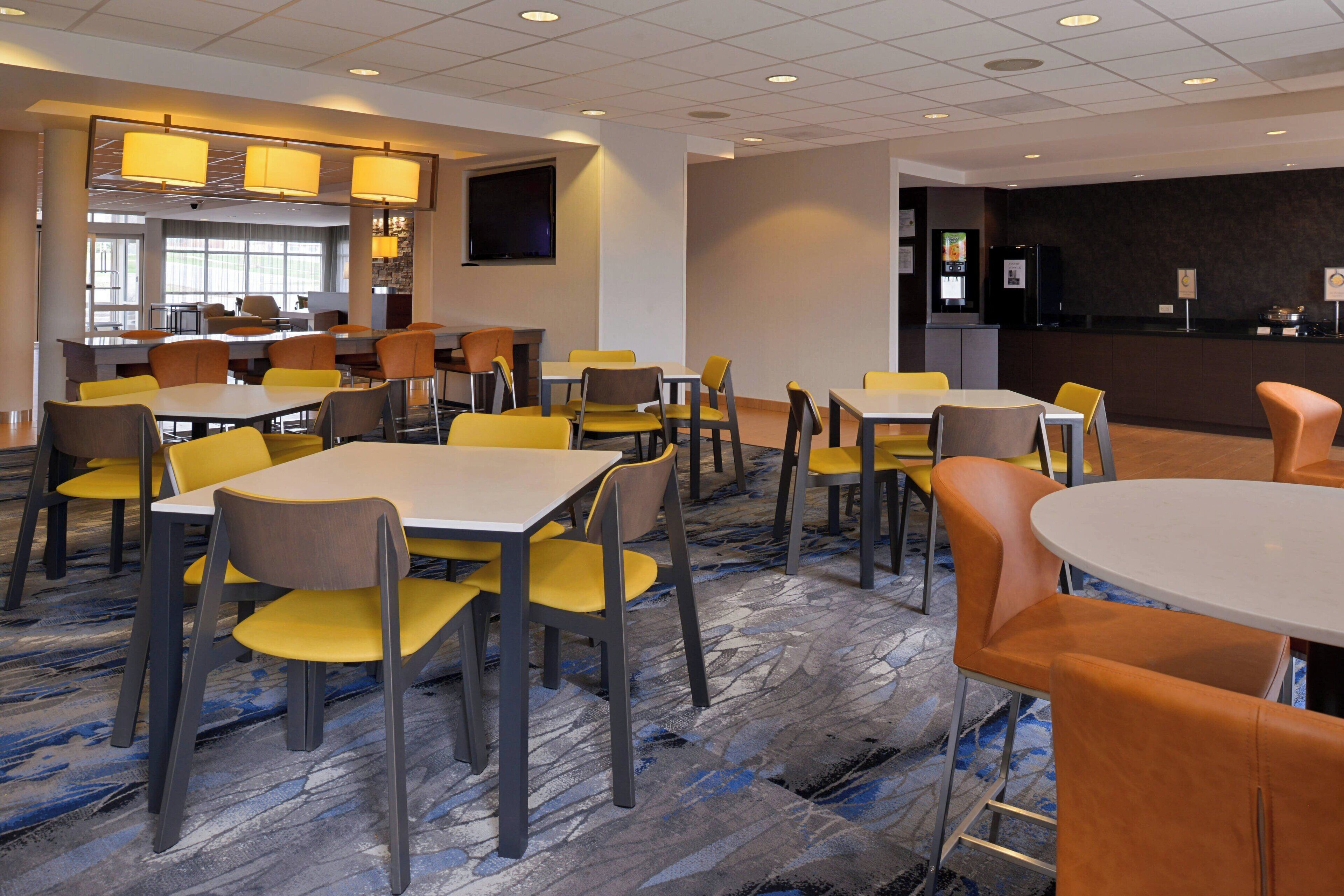 A hotel lobby with multiple dining tables and chairs, a reception desk, and a large screen on the wall.