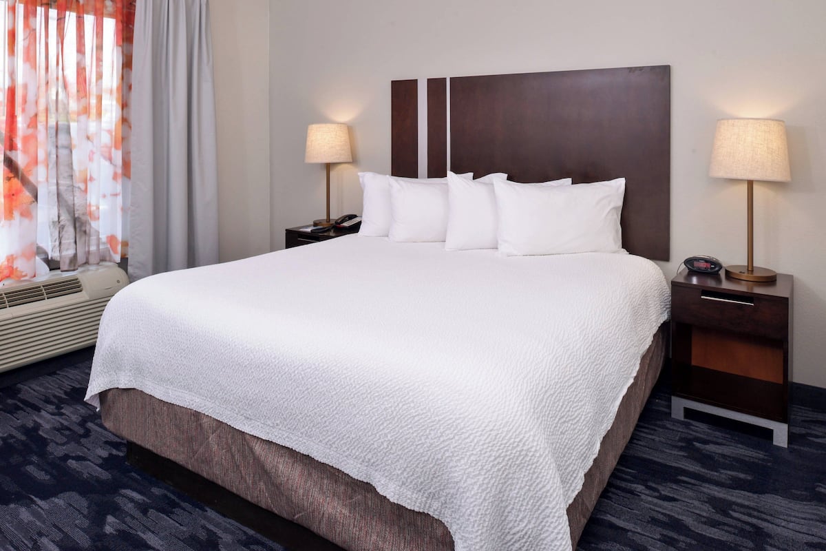 A hotel room with a large bed, two bedside tables with lamps, a window with curtains, and an air conditioning unit.