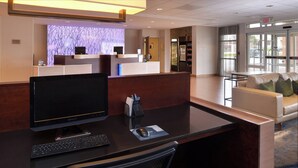 A reception desk with a computer, a keyboard, and a mouse. There is a reception area with a counter, a refrigerator, and a glass door leading outside. A white sofa is visible in the background. - Fairfield Inn & Suites Cedar Rapids (Cedar Rapids)