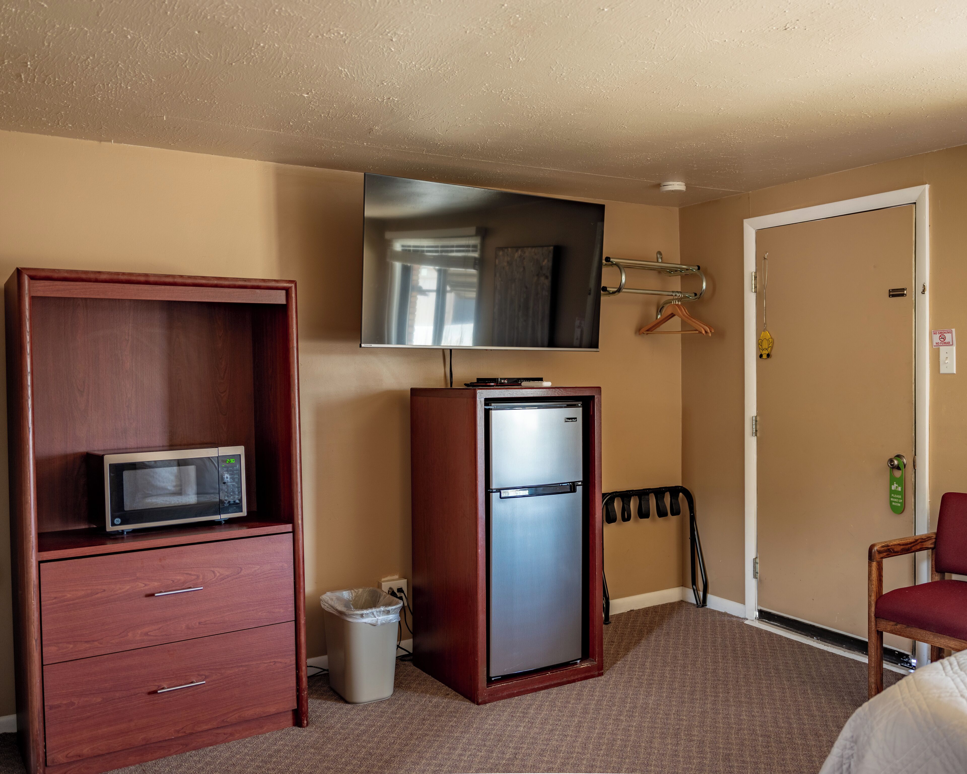 A hotel room with a wooden cabinet, a microwave, a refrigerator, a trash can, a TV mounted on the wall, a coat rack, a door, and a chair.