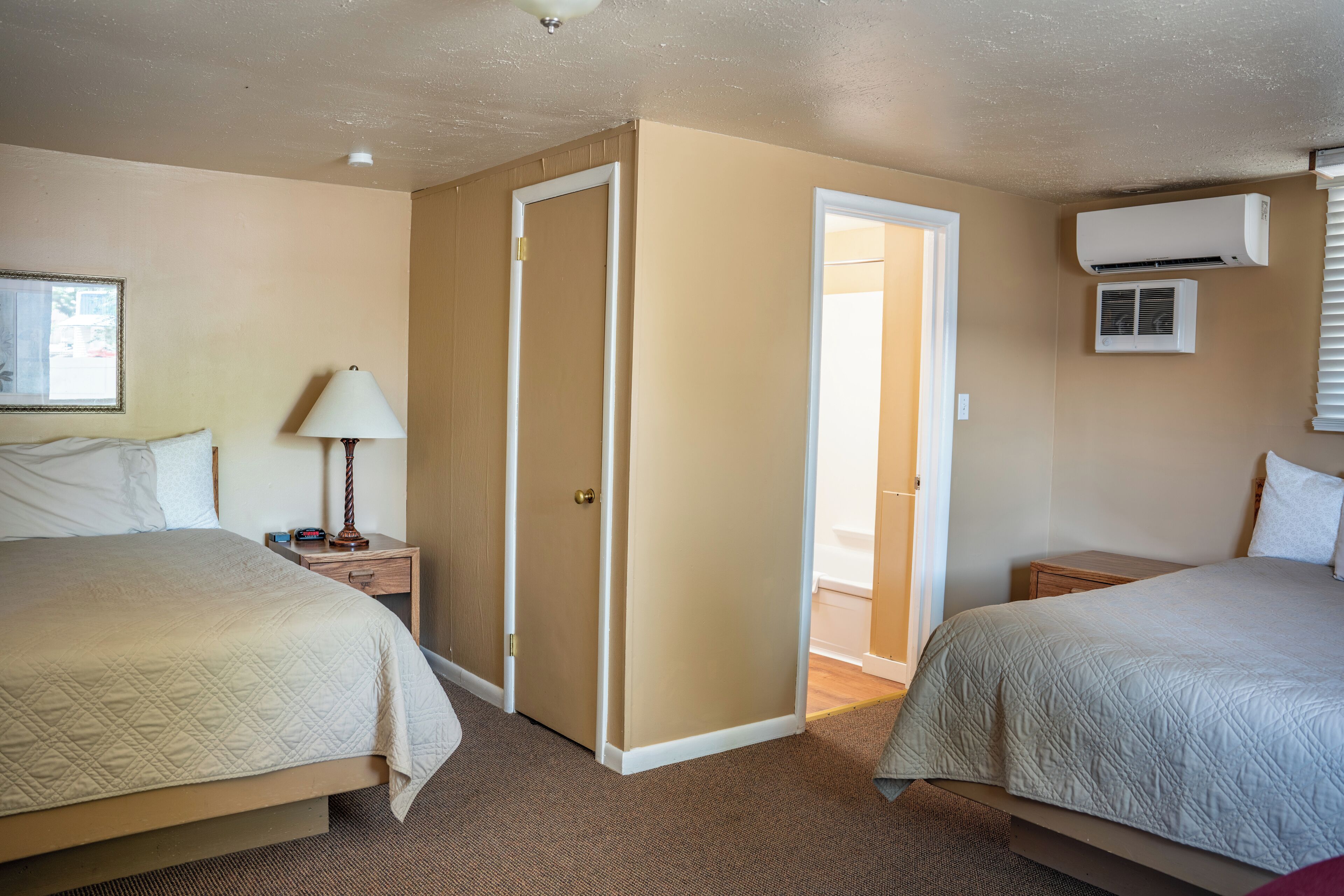 A hotel room with two beds, a bedside table with a lamp, an air conditioning unit, and a door leading to another room.