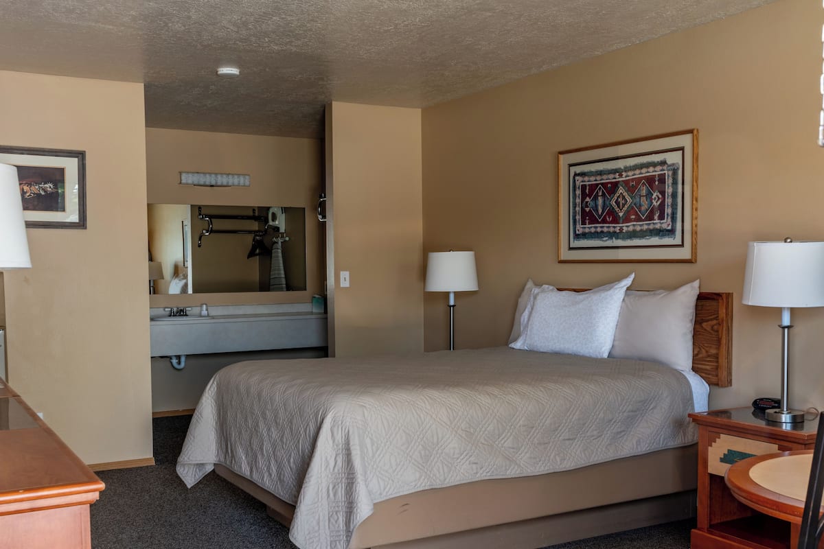 Quarto de hotel com cama, mesas de cabeceira, escrivaninha, cadeira, abajur, espelho e quadro na parede.