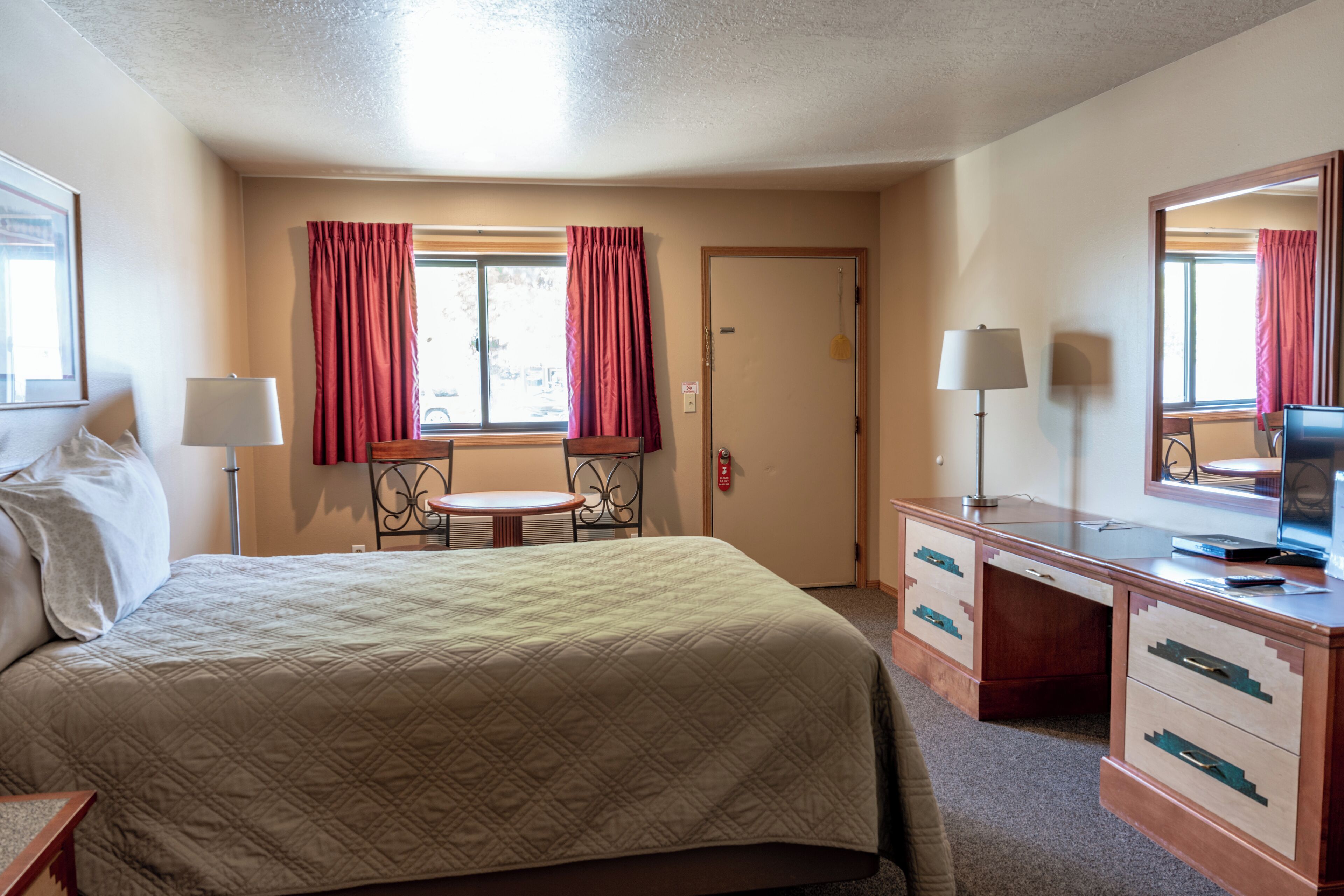 Quarto de hotel com cama, escrivaninha, cadeira, abajur e janela com cortinas.