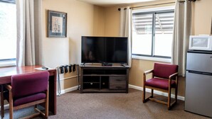 A compact hotel room with a TV on a wooden stand, a small refrigerator, a desk with a chair, and a window with curtains. - Oregon Trail Inn (Buhl)