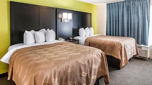 A hotel room with two beds, a large headboard, a nightstand, a wall-mounted lamp, and a window with curtains.