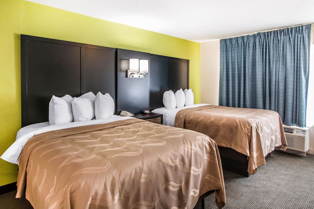 A hotel room with two beds, a large headboard, a nightstand, a wall-mounted lamp, and a window with curtains.