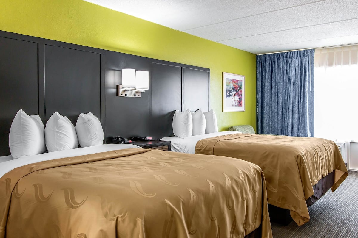 A hotel room with two beds, a desk, a nightstand, and a window with curtains.