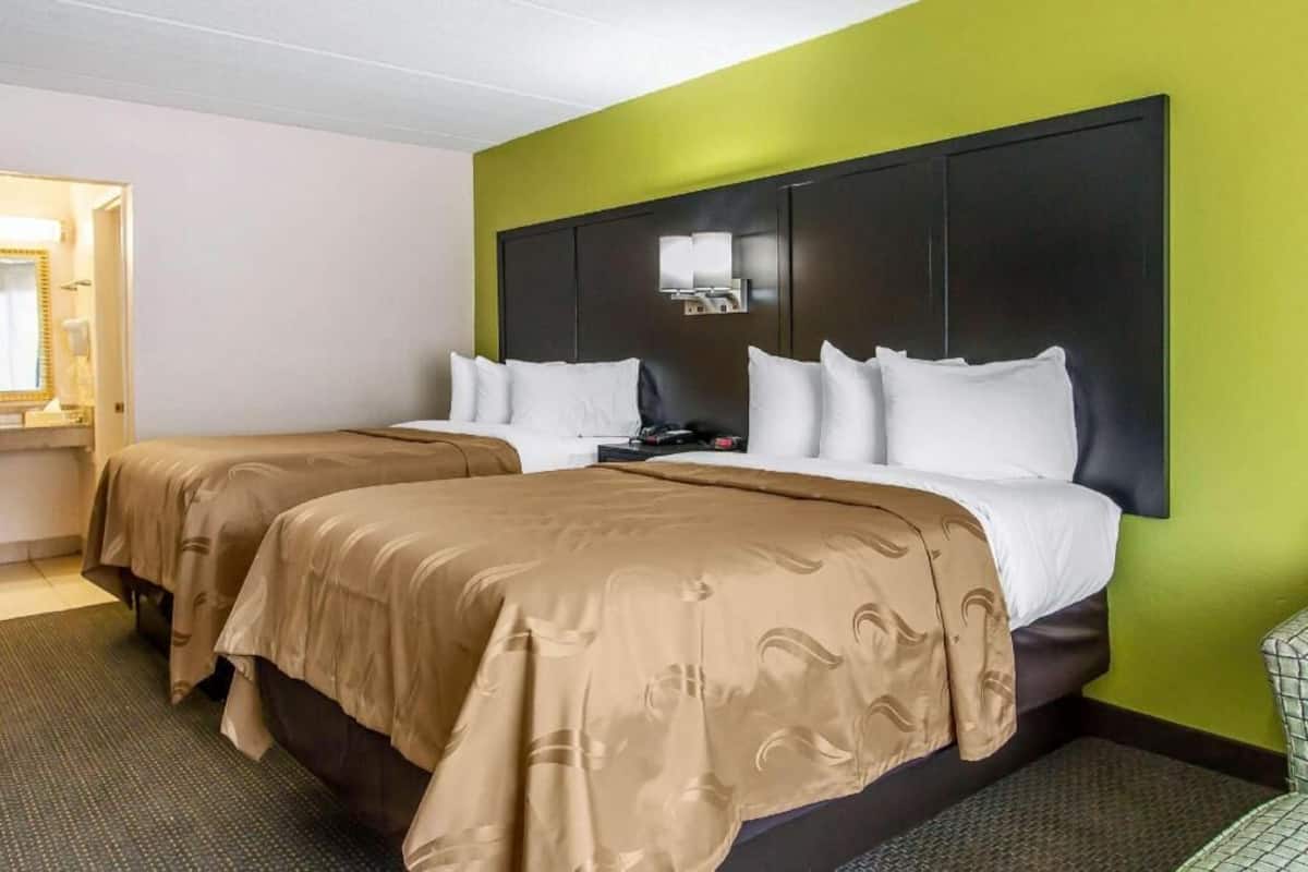 A hotel room with two beds, a headboard with a mirror, and a small table with a lamp.