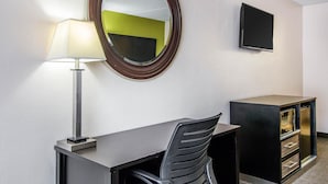 A hotel room with a desk, chair, lamp, mirror, TV, and a cabinet.