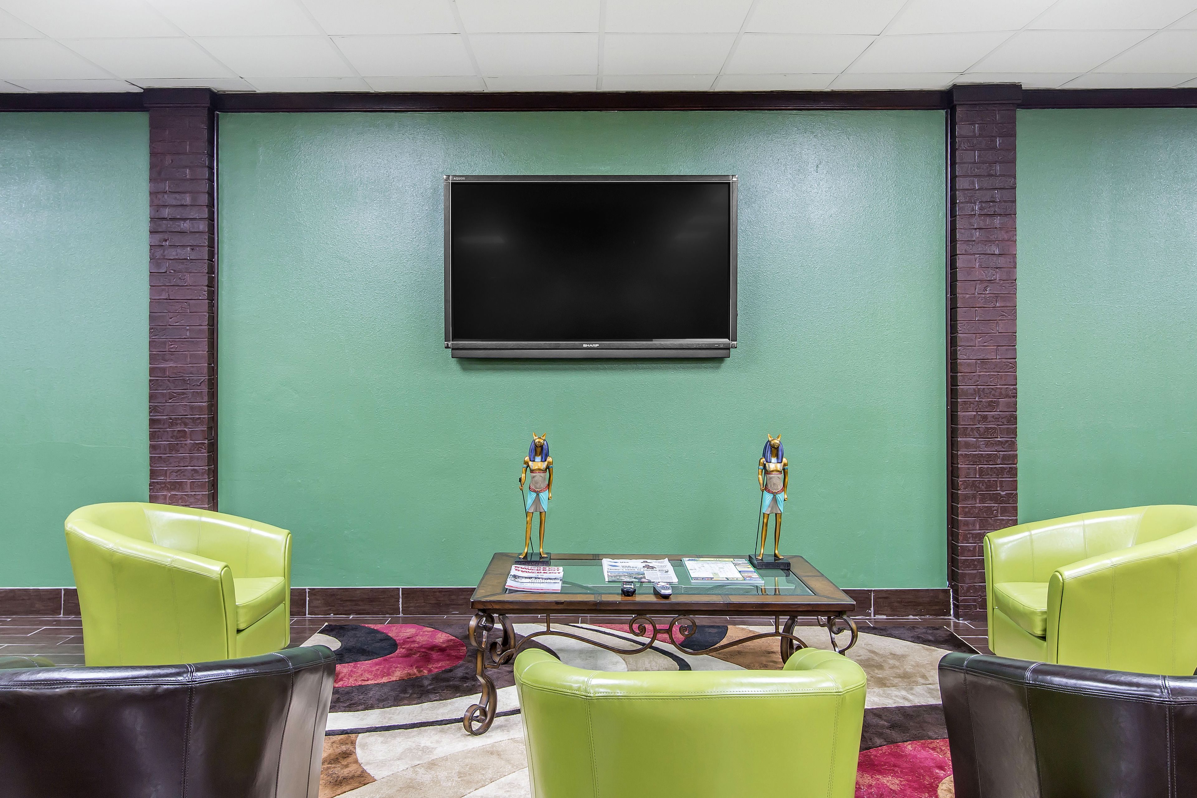 A conference room with a large flat-screen TV mounted on the wall, two green chairs, a glass-top table with papers, and decorative statues.