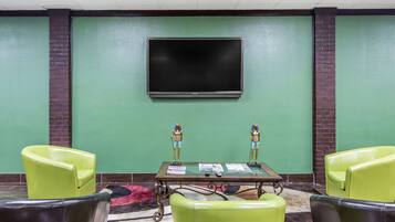 Un salón de conferencias con una televisión de pantalla plana grande montada en la pared, dos sillas verdes, una mesa con cubierta de vidrio y papeles, y estatuas decorativas.