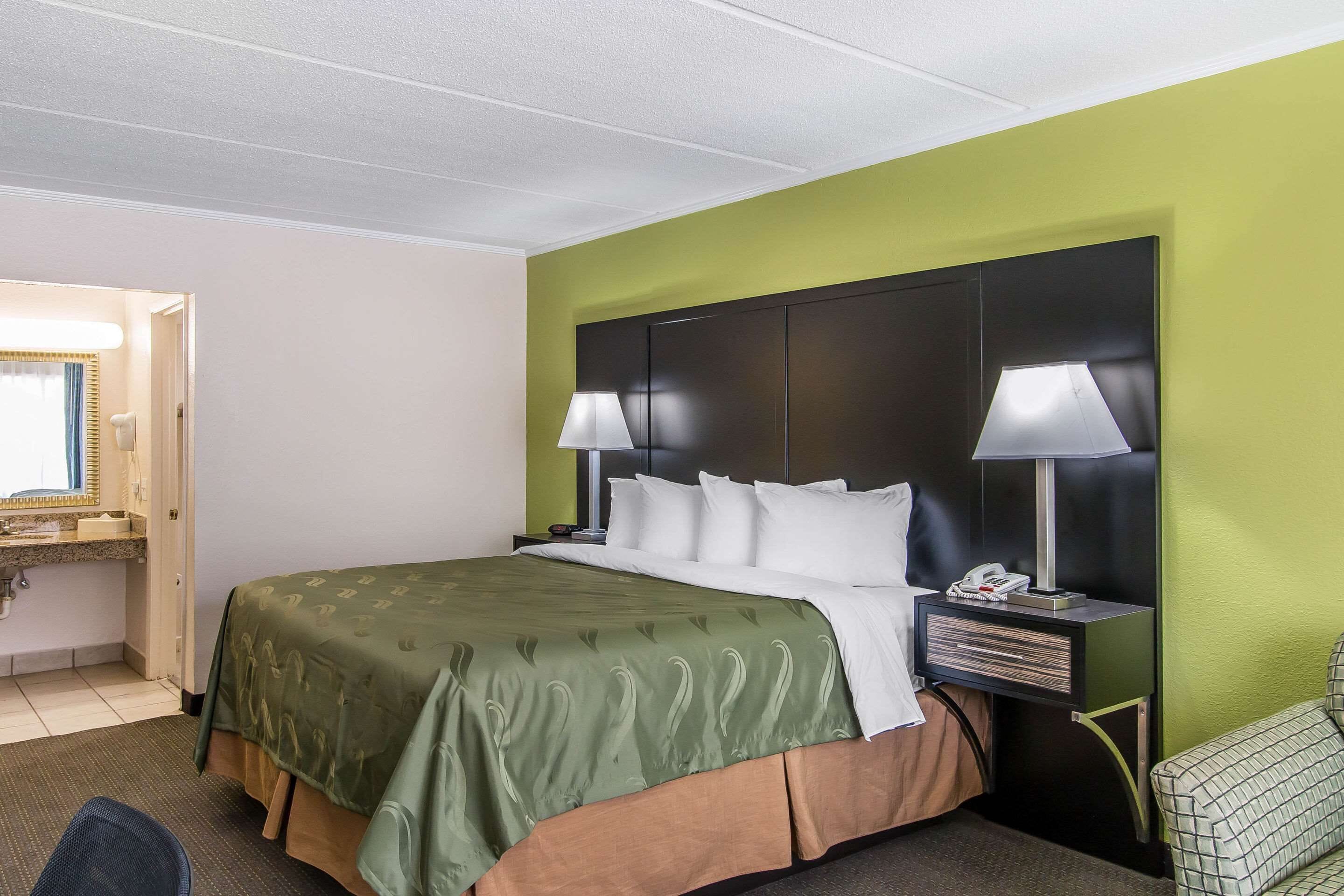 A hotel room with a large bed, two bedside lamps, a nightstand, a green wall, and a small chair.