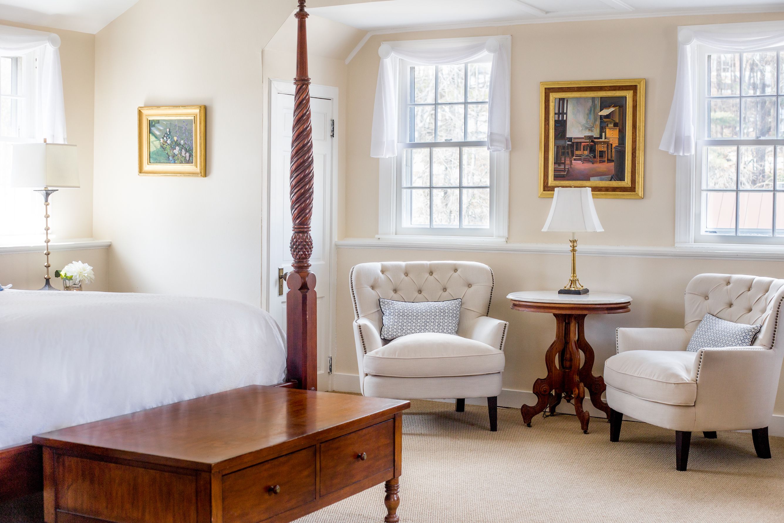 A bedroom with a four-poster bed, two armchairs, a round wooden table, and two framed pictures on the walls.