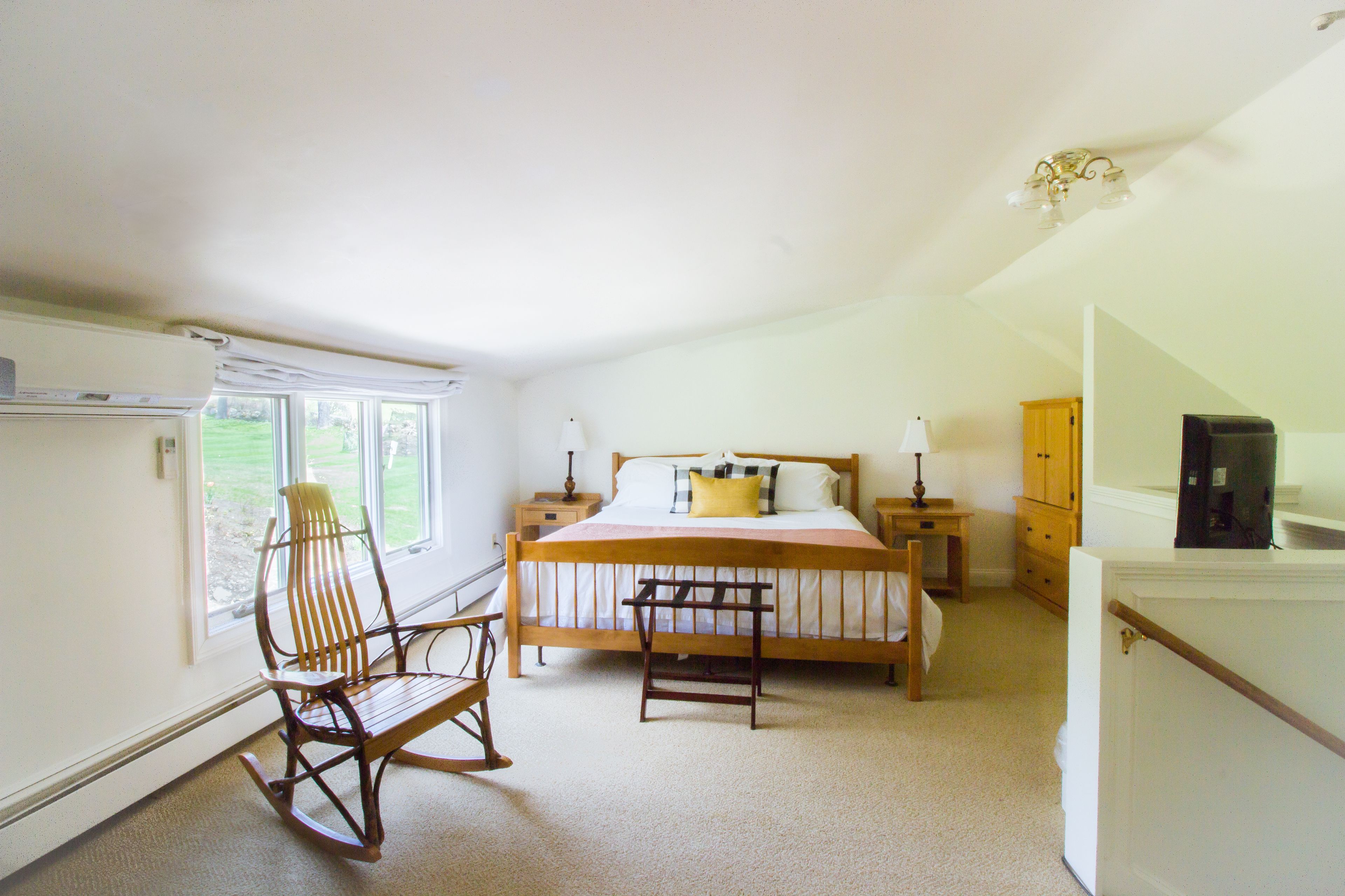 A bedroom with a bed, a rocking chair, a nightstand, a dresser, and a window with a view of greenery.