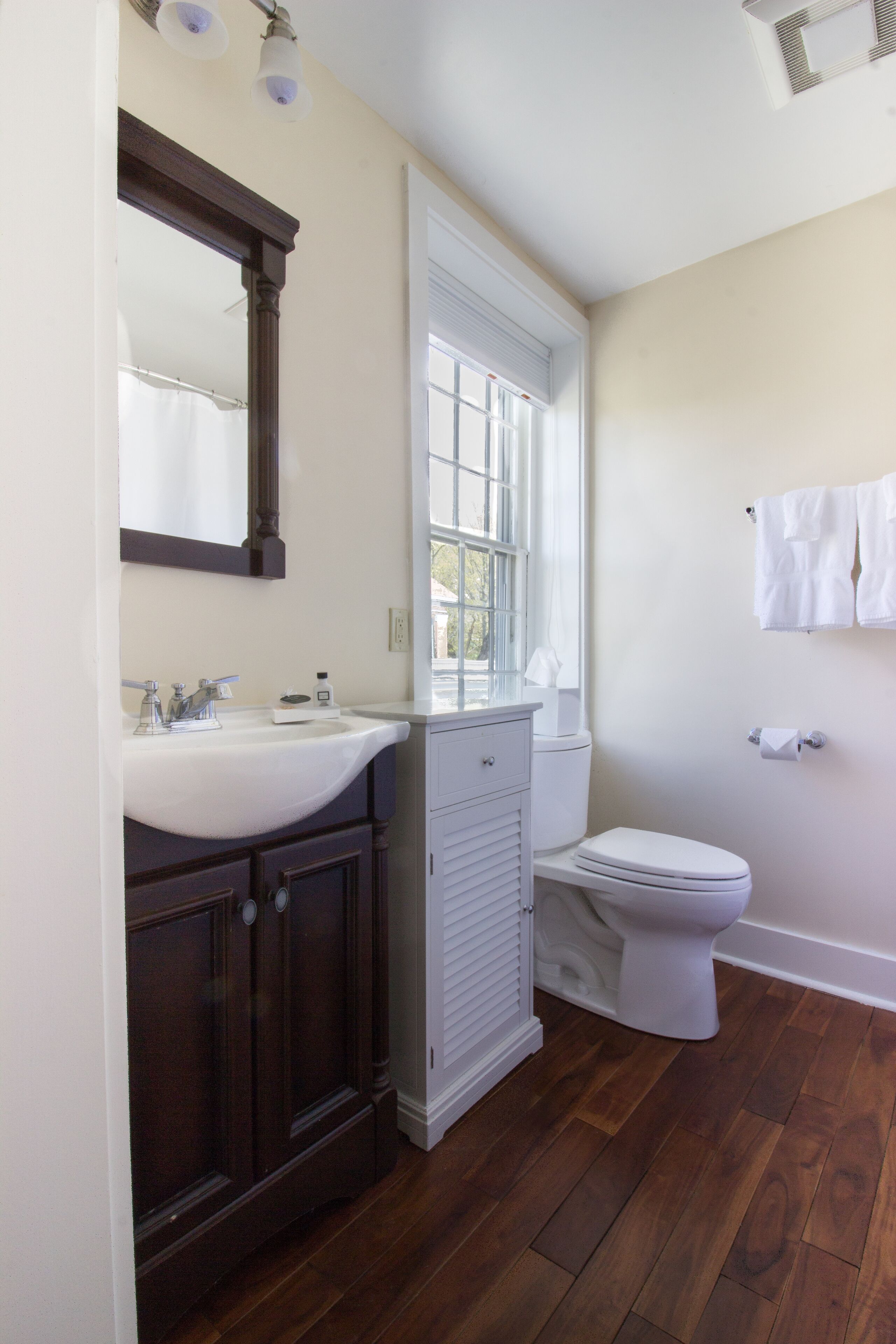 Une salle de bain avec un lavabo blanc, des meubles en bois foncé, des toilettes blanches et une fenêtre avec des stores.