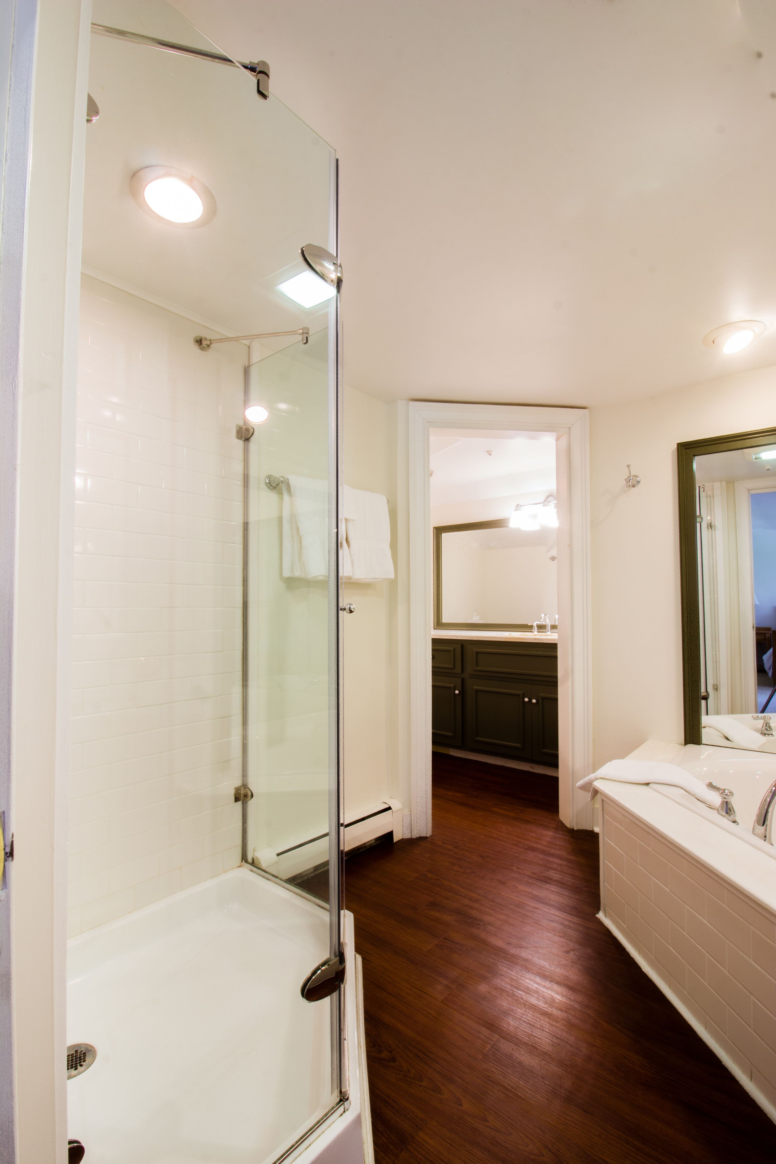 Un baño moderno con ducha de vidrio, piso de madera y lavamanos blanco con espejo.