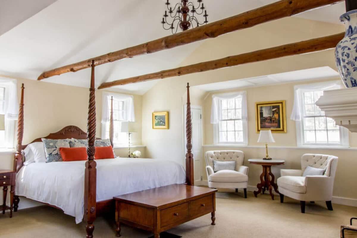 A four-poster bed with a wooden headboard, a bedside table, and two armchairs in a room with large windows and a chandelier.