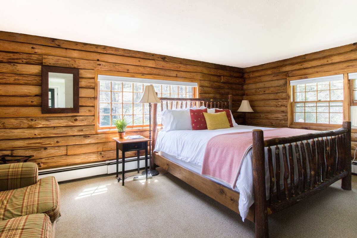 A log cabin bedroom with a wooden bed, a nightstand, a chair, and a window with a view of trees.