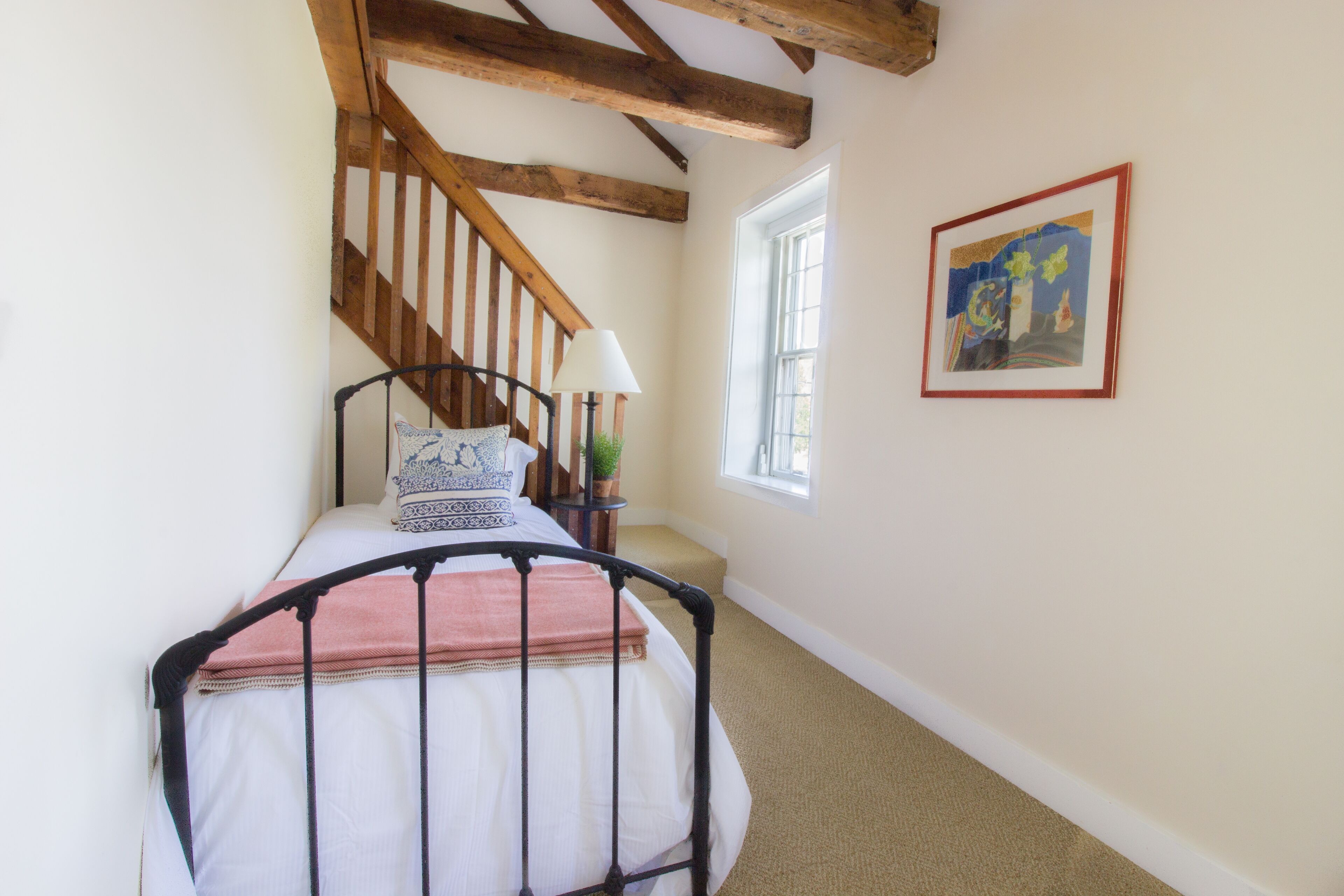 A bedroom with a bed, wooden staircase, a window, and a framed picture on the wall.