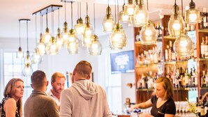 Uma área de bar com banquetas de madeira, luminárias pendentes e um bartender no balcão.