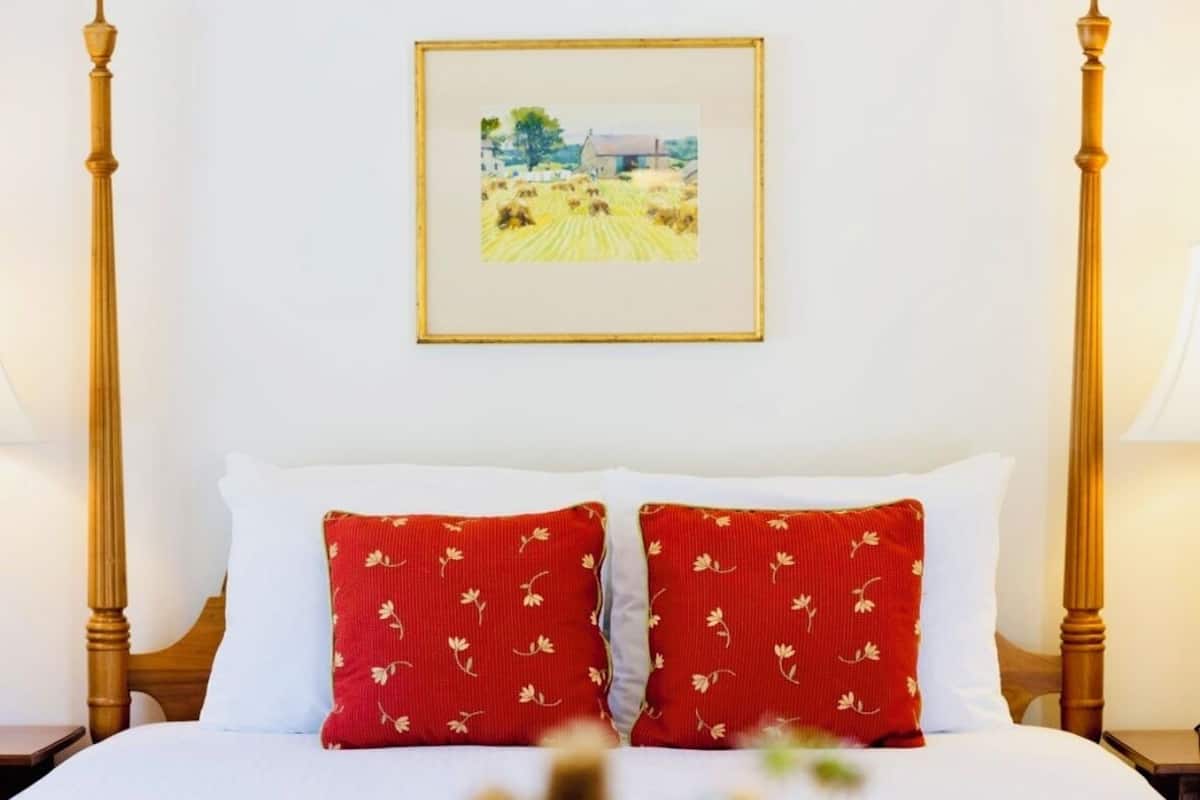 A neatly made bed with white and red pillows, a framed picture on the wall, and a vase with flowers on the nightstand.