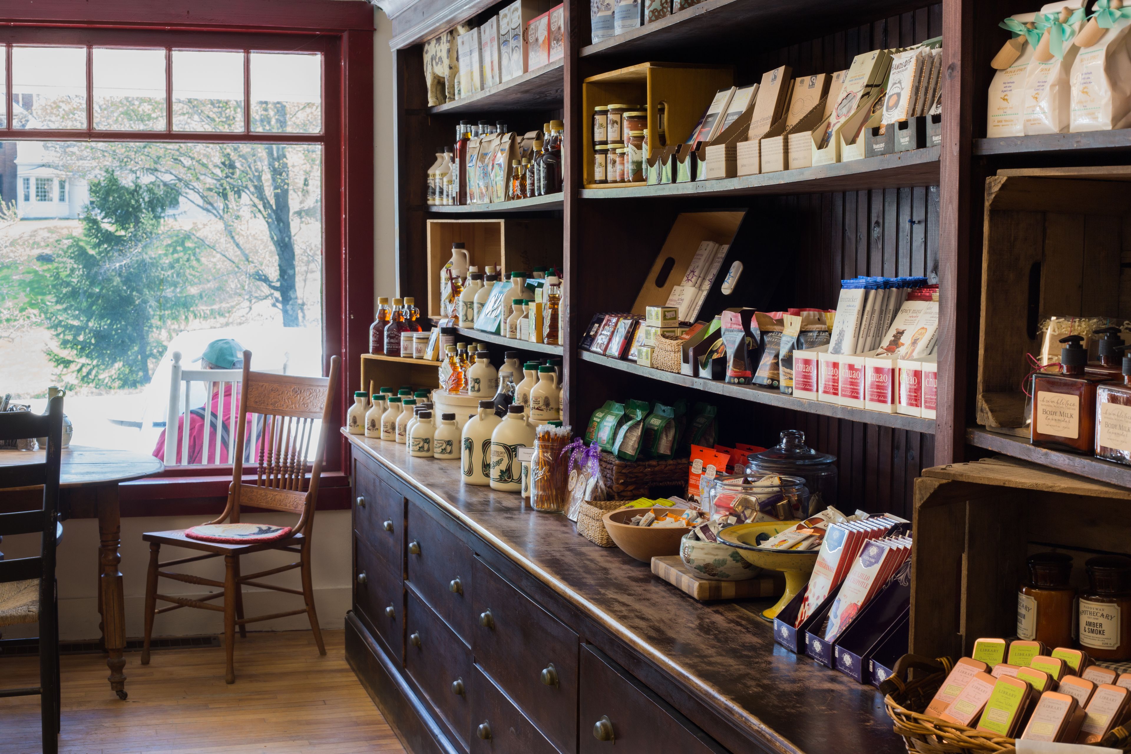 Un intérieur rustique avec des étagères en bois remplies de divers produits alimentaires et une chaise en bois près d’une fenêtre.