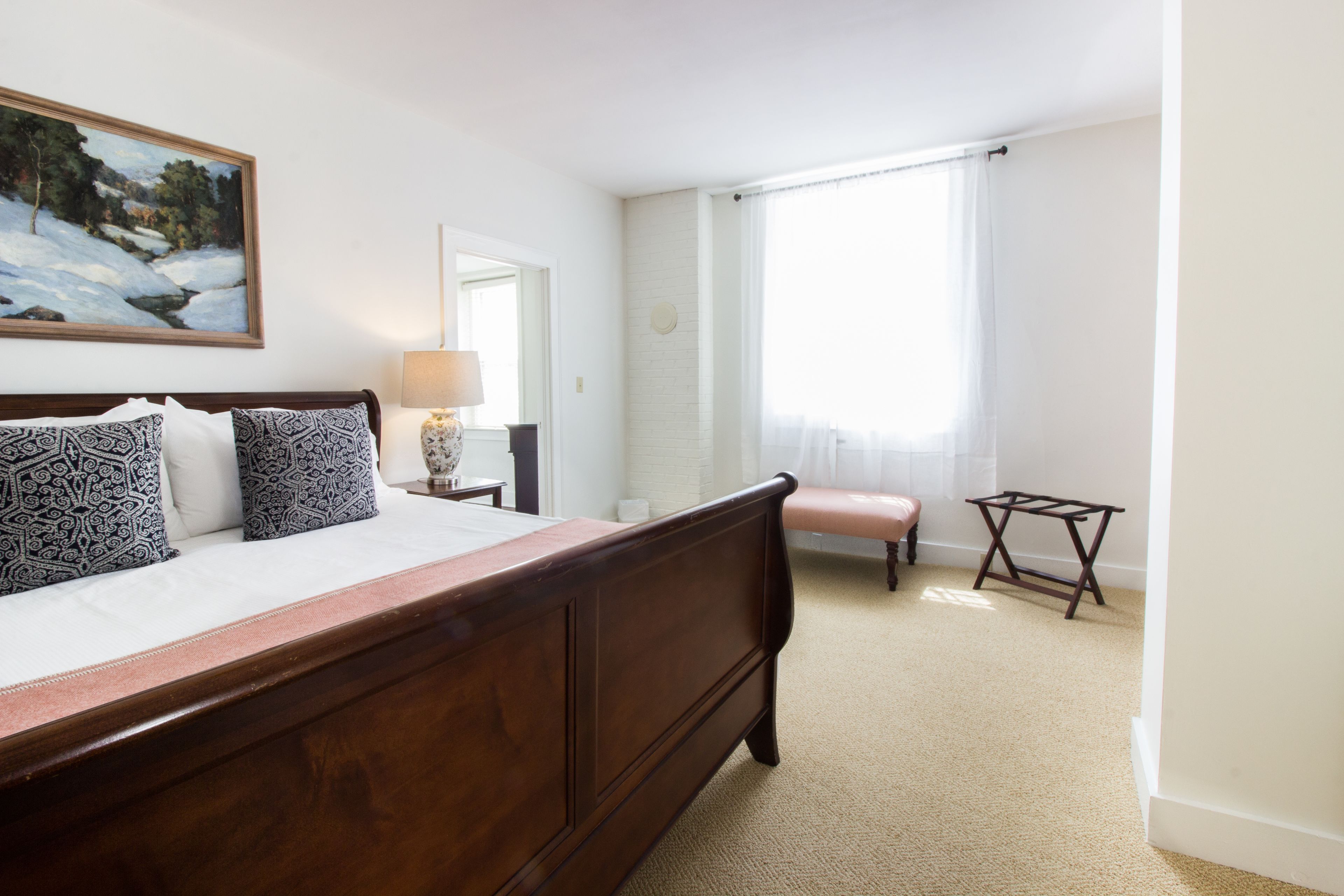 A bedroom with a wooden bed, patterned pillows, a painting, a lamp, a window with curtains, and a small table.
