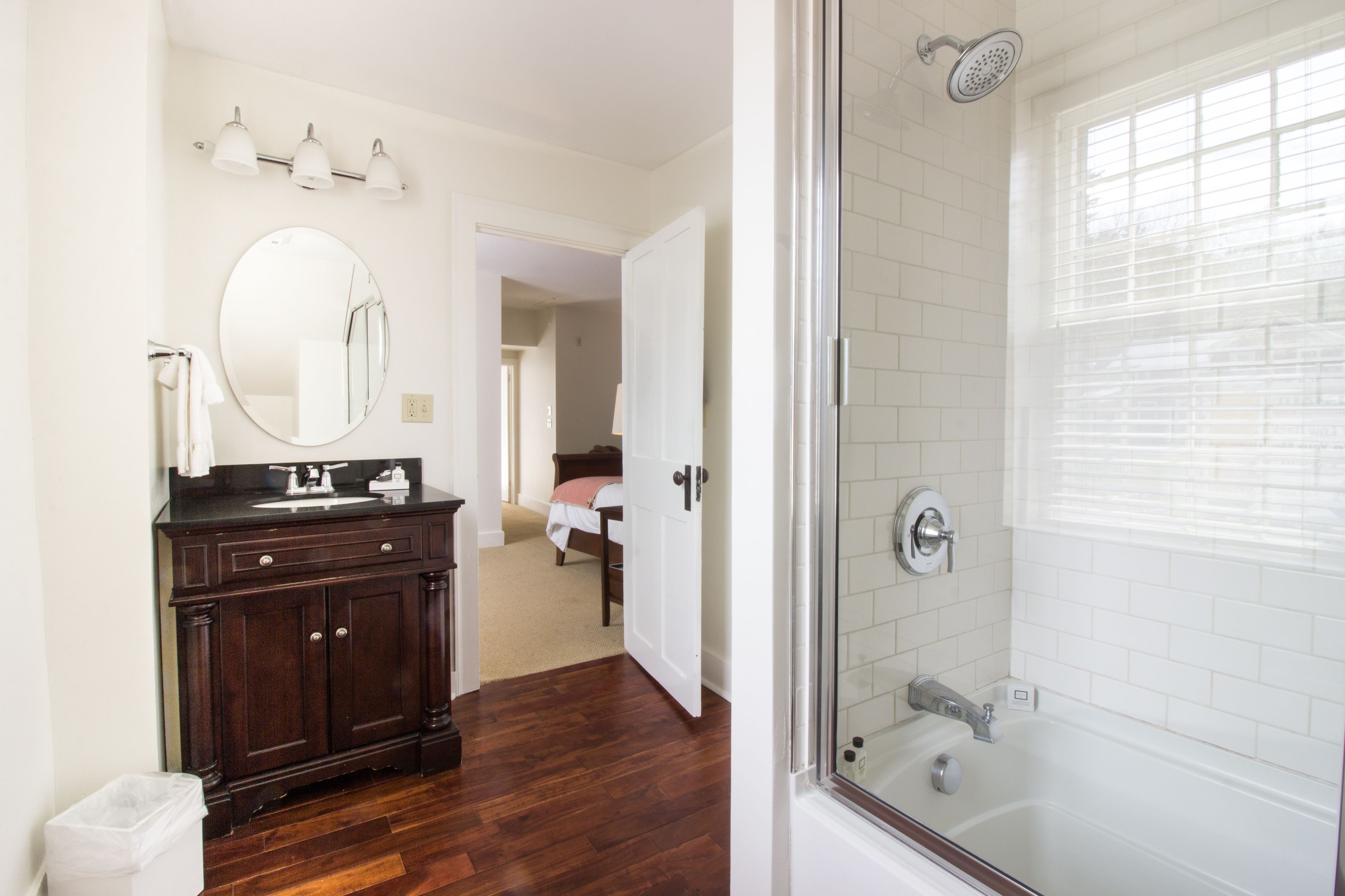Un baño con tocador de madera oscura, espejo redondo, lavabo blanco y ducha con cerramiento de vidrio.