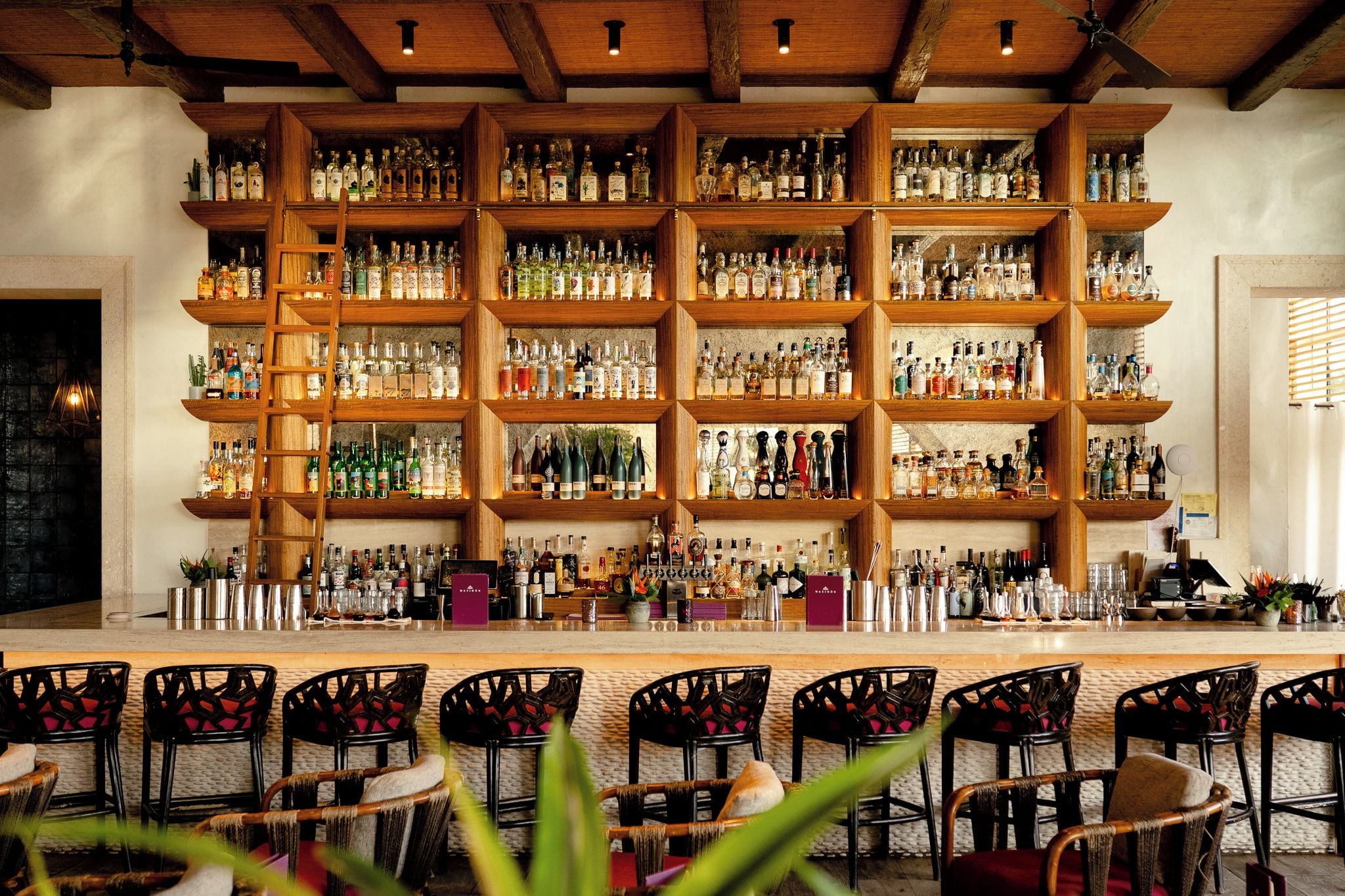 Un espace bar doté de plusieurs étagères remplies de bouteilles, d’un comptoir avec des ustensiles de bar et de chaises en osier.