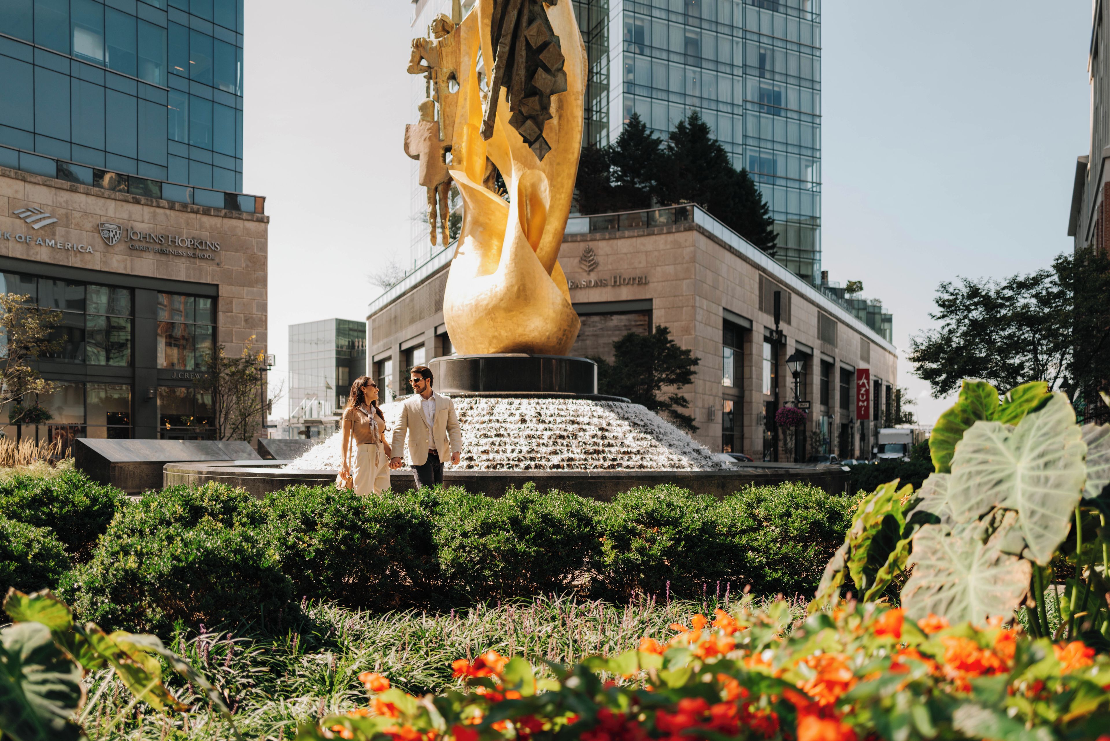 Un couple se promène devant une grande sculpture dorée, dans un espace aménagé avec des bâtiments en arrière-plan.