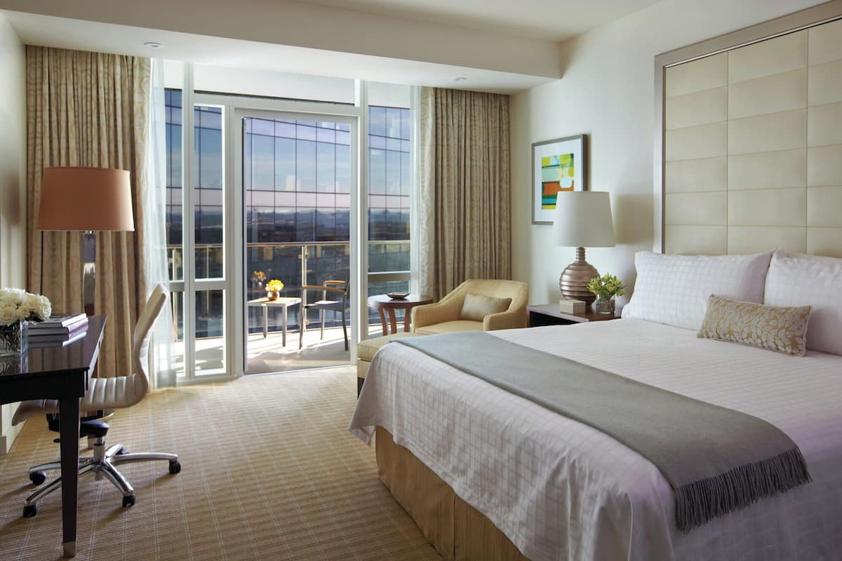 A hotel room with a large bed, a desk with a chair, a small table with a lamp, and a view through the glass doors.