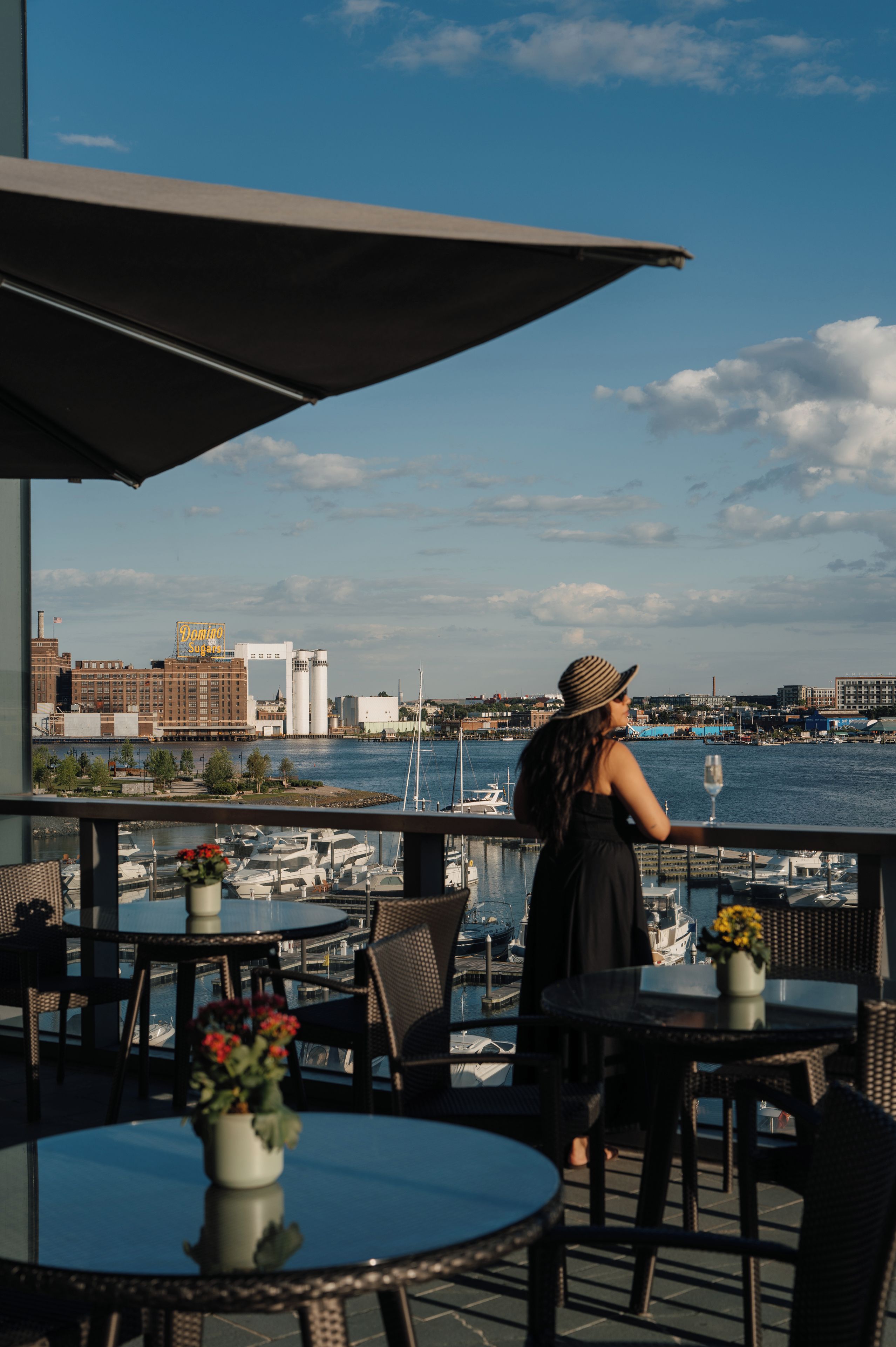 Una mujer de pie junto a una mesa, con vista a un puerto deportivo y edificios al otro lado del agua.