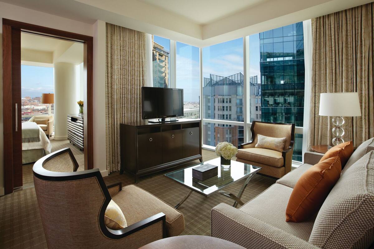 A modern hotel room with a large window, a flat-screen TV, a sofa, a coffee table, and a view of the cityscape.