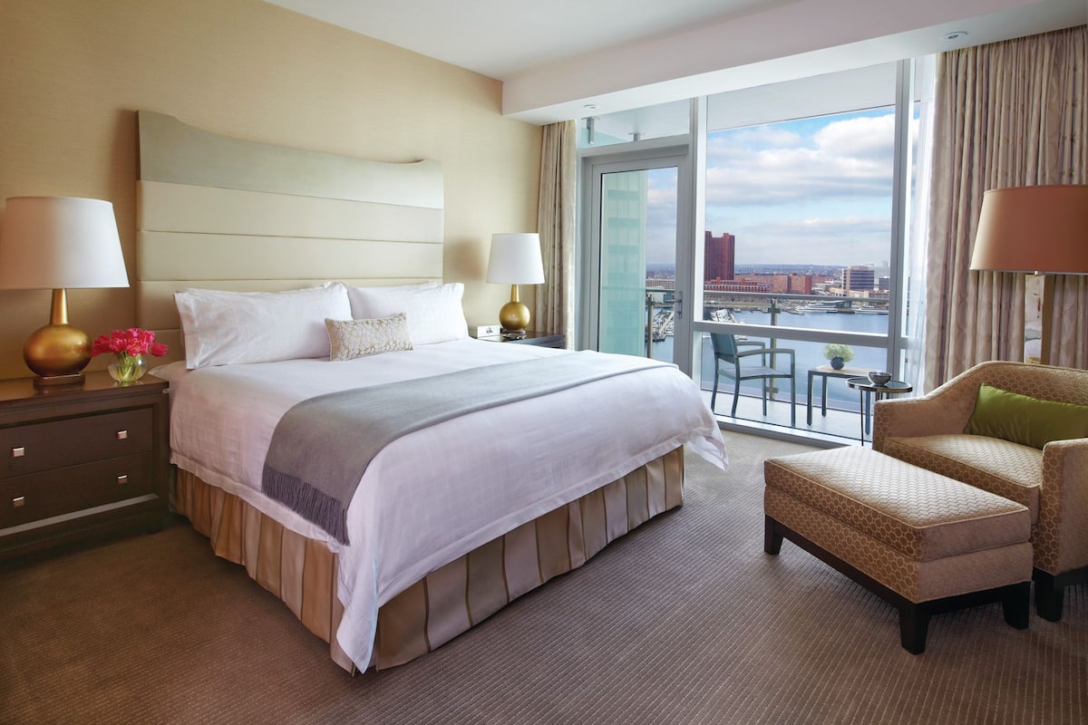 A hotel room with a large bed, two bedside lamps, a chair, a small table, and a view of the city through a large window.