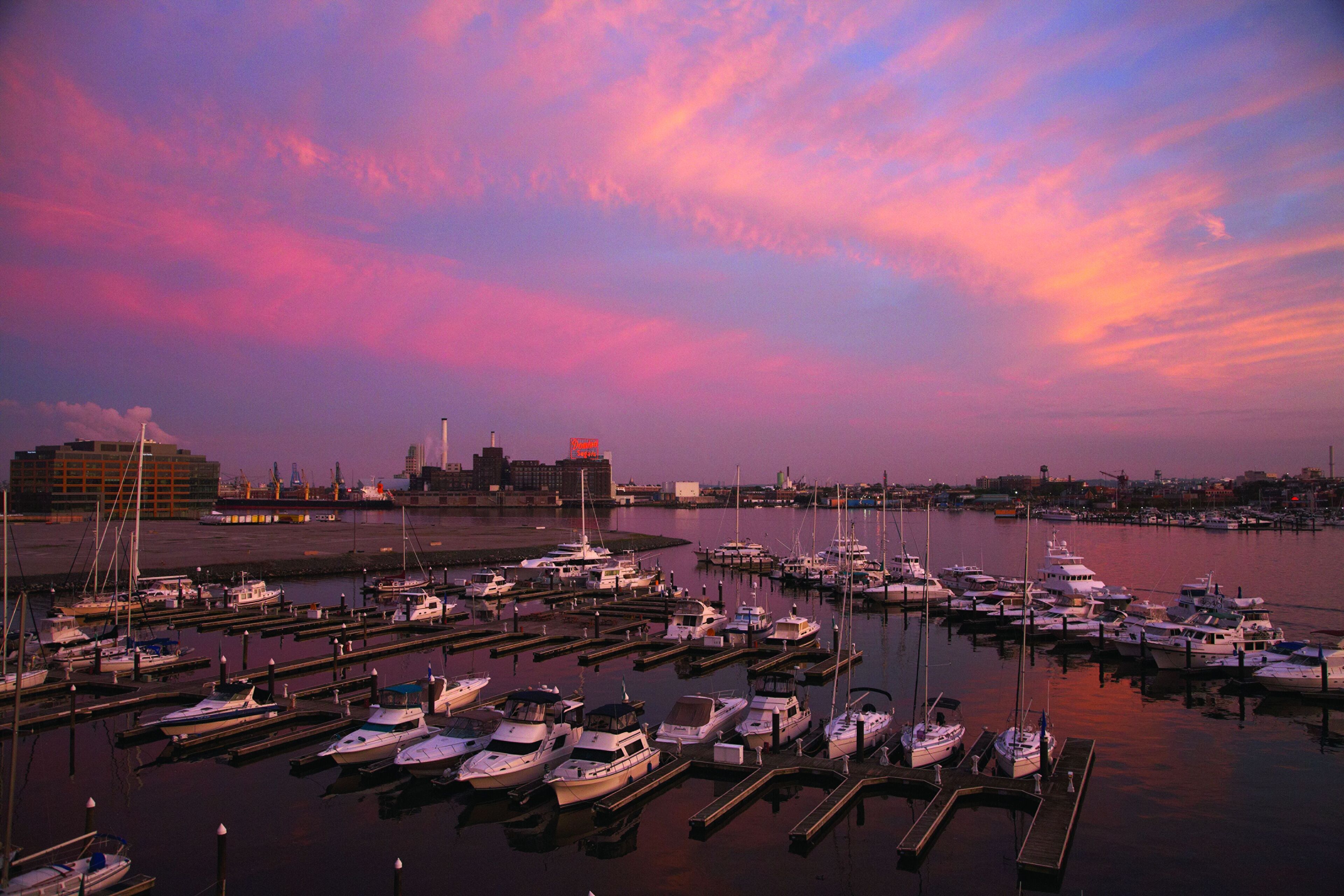 A marina with boats docked at piers, a city skyline in the background, and a sunset sky.