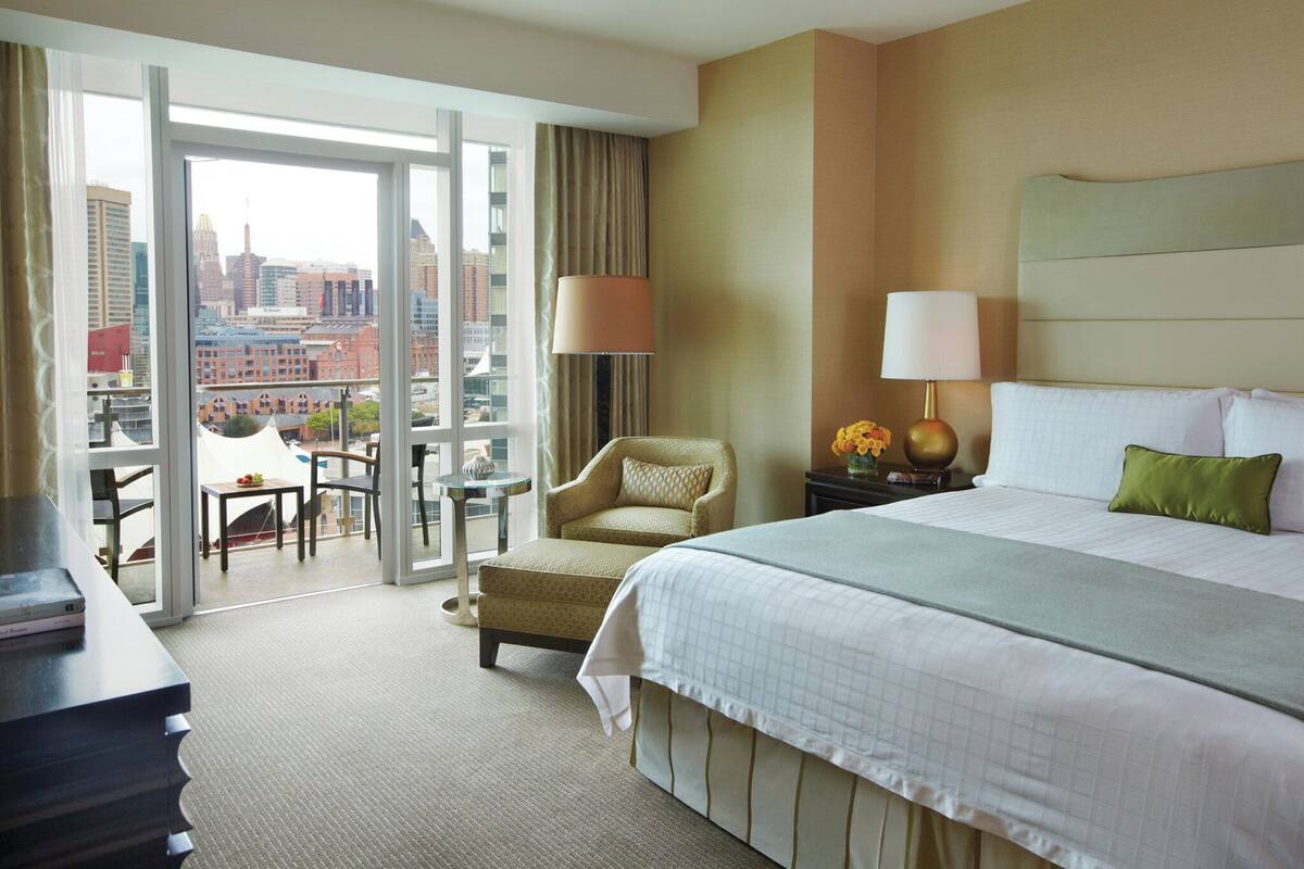 A hotel room with a large bed, a chair, a table, and a view of the cityscape through the window.
