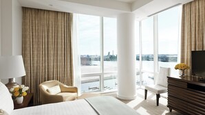 Une chambre d’hôtel moderne dotée d’un grand lit, de deux fauteuils, d’une télévision et offrant une vue sur le paysage urbain grâce à de grandes fenêtres.