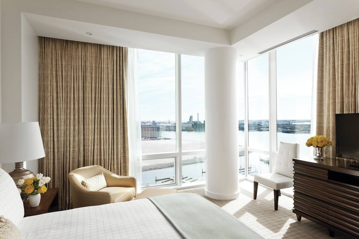 A modern hotel room with a large bed, two chairs, a TV, and a view of a cityscape through large windows.