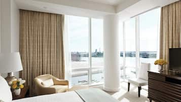Habitación de hotel moderna con una cama grande, dos sillones, un televisor y vistas a un paisaje urbano a través de amplias ventanas.