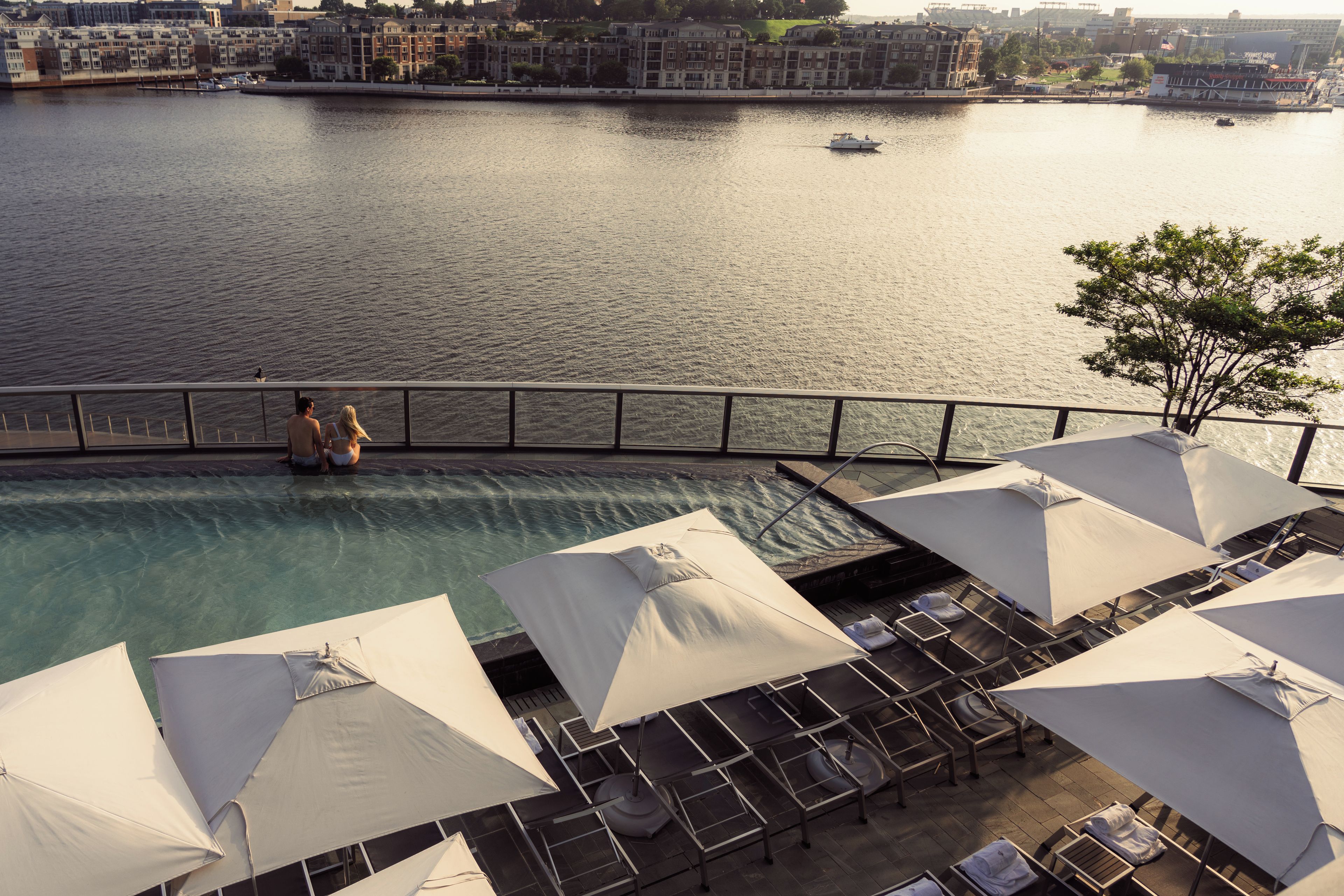 A poolside area with lounge chairs and umbrellas, overlooking a calm body of water with boats.