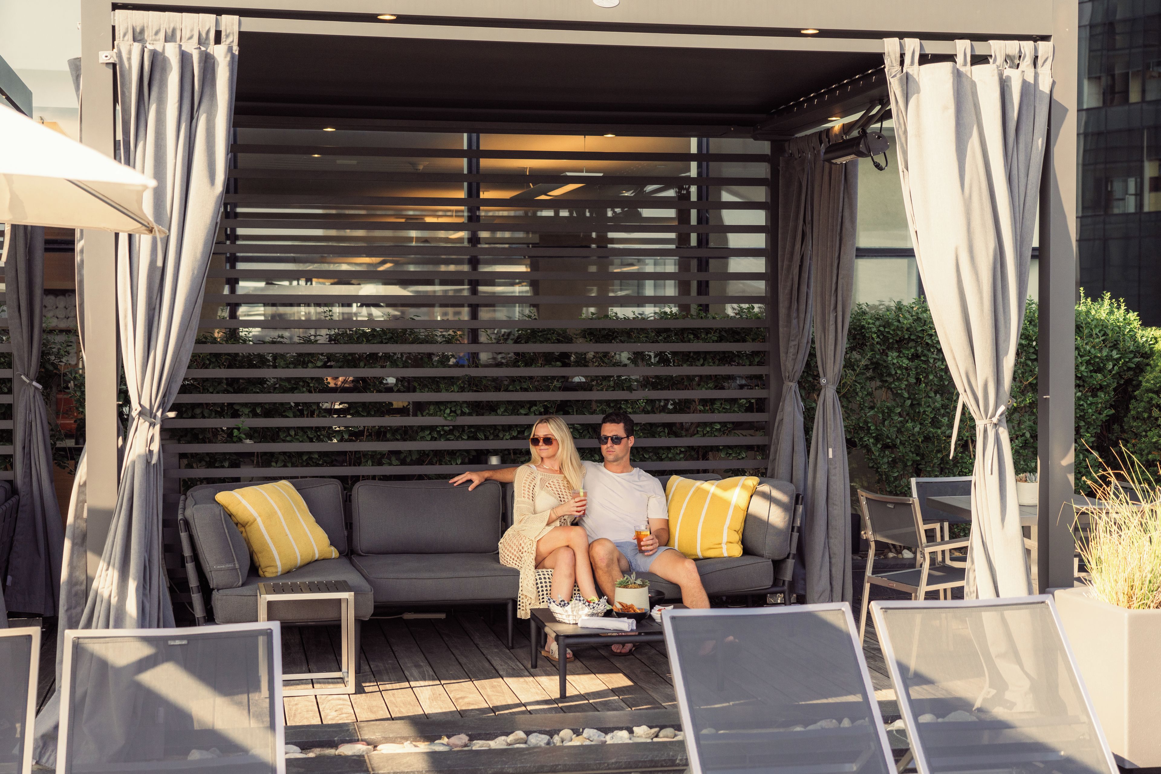 Un couple est assis sur un canapé gris agrémenté de coussins, près d’une petite table et d’une plante en pot, dans un décor extérieur moderne surmonté d’une pergola.
