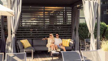 Un couple est assis sur un canapé gris agrémenté de coussins, près d’une petite table et d’une plante en pot, dans un décor extérieur moderne surmonté d’une pergola.